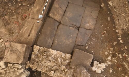 Monumentaler Eingang mit Sandsteinschwelle (bei der Messlatte und Fototafel) und Plattenbelag. Der Sandsteinquader unten rechts diente wohl als Basis für eine Säule. © Augusta Raurica